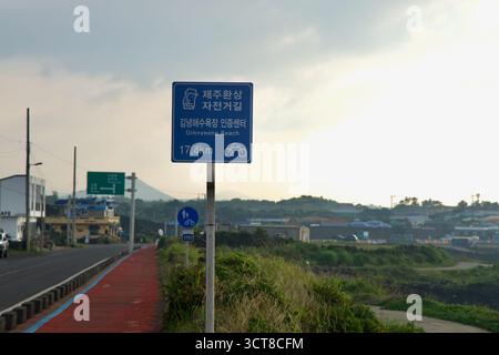 Un cartello blu sulla pista costiera punta verso il centro di certificazione di Gimnyeong Beach, con il sentiero pavimentato di rosso e i tetti del villaggio che si estendono su ALE Foto Stock
