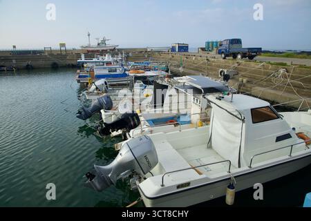 Piccole barche da pesca con motori fuoribordo sono ormeggiate all'interno del bacino di cemento del porto di Hado sull'isola di Jeju, con camion da lavoro e attrezzature portuali lungo la banchina Foto Stock