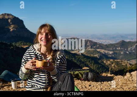 Una donna allegra in una felpa a righe si siede su un terreno roccioso, tenendo una tazza di caffè con una vasta catena montuosa e un cielo limpido dietro di lei, zaino vicino Foto Stock