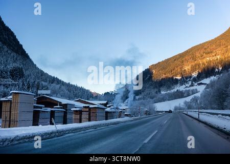 La tranquilla strada invernale si snoda attraverso una valle di montagna accanto a un cortile di legname. Il fumo sorge dalle case del villaggio mentre la luce del sole dorata riscalda la foresta Foto Stock