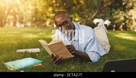 Felice ragazzo nero studente in occhiali lettura libro, la preparazione per gli esami e godere di studiare all'aperto nel parco Foto Stock