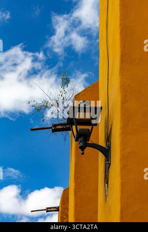 Un'immagine ad angolo basso di un lampione nero attaccato a un edificio giallo con un piccolo albero che cresce fuori dal muro accanto, adagiato contro un cielo blu Foto Stock