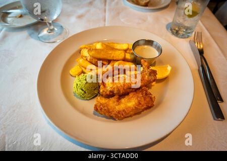 Classico fish and chips con merluzzo pastella dorato, patatine fritte croccanti, vivaci piselli e spicchi di limone, serviti con un lato di cremosa salsa tartare Foto Stock