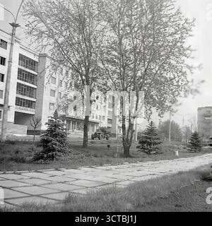 Una tranquilla strada residenziale a Sloviansk, RSS Ucraina, durante gli anni '1960 La fotografia mostra la nuova architettura sovietica dell'epoca, con condomini in mattoni a cinque piani caratterizzati da grandi logge di vetro. Il primo piano mostra un'area recentemente paesaggistica con giovani abeti e una passerella fatta di lastre di cemento. Un dettaglio insolito e sorprendente è l'auto da ricognizione corazzata militare, probabilmente un BRDM-1, parcheggiata casualmente sul prato tra gli alberi. Foto Stock