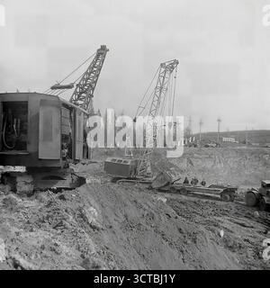 Un progetto di movimento terra e costruzione su larga scala in corso nell'area di Sloviansk, RSS Ucraina, durante gli anni '1960 Questa fotografia d'archivio cattura la potenza grezza dei macchinari pesanti sovietici al lavoro. Le enormi gru cingolate dominano la vasta fossa di scavo, sollevando e spostando terra e materiali per preparare le fondamenta per un nuovo progetto di fabbrica o infrastruttura. I lavoratori possono essere visti indeboliti dalla portata delle attrezzature, evidenziando l'ambizione industriale dell'epoca. Questa immagine è una testimonianza dei progetti trasformativi che hanno rimodellato il paesaggio della regione del Donbas Foto Stock