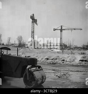 Un aspro paesaggio invernale in un cantiere negli anni '1960 di Sloviansk, RSS Ucraina. La fotografia cattura la fase preparatoria di un nuovo sviluppo abitativo durante il boom sovietico della costruzione di massa. Due grandi gru a torre, indispensabili per la costruzione di condomini "Khrushchyovka", sono pronte all'azione contro un cielo grigio. In primo piano, un rullo innevato attende su una strada incompiuta. Questa immagine atmosferica simboleggia la scala industriale e l'ambizione dell'epoca, mostrando i macchinari che hanno trasformato lo skyline della città e creato nuove case. Foto Stock