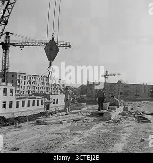 Una vista da un cantiere nel nuovo quartiere Pushkin Boulevard di Sloviansk, RSS Ucraina, durante gli anni '1960 La fotografia cattura i costruttori al lavoro su una nuova struttura, con il massiccio gancio di una gru a torre appesa sopra la testa. Sullo sfondo, file di appartamenti "Khrushchyovka" recentemente completati rappresentano una testimonianza del boom edilizio sovietico. Questa immagine fornisce un autentico sguardo dietro le quinte del processo di costruzione, evidenziando il lavoro manuale che ha costruito i nuovi microquartieri della città. Foto Stock