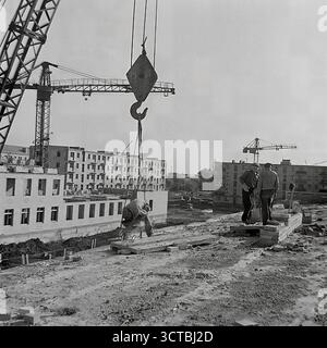 Una vista da un cantiere nel nuovo quartiere Pushkin Boulevard di Sloviansk, RSS Ucraina, durante gli anni '1960 La fotografia cattura i costruttori al lavoro su una nuova struttura, con il massiccio gancio di una gru a torre appesa sopra la testa. Sullo sfondo, file di appartamenti "Khrushchyovka" recentemente completati rappresentano una testimonianza del boom edilizio sovietico. Questa immagine fornisce un autentico sguardo dietro le quinte del processo di costruzione, evidenziando il lavoro manuale che ha costruito i nuovi microquartieri della città. Foto Stock