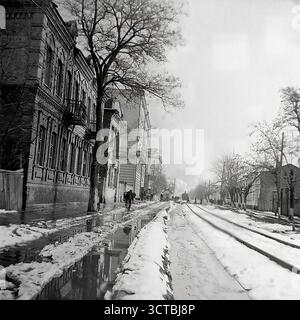 Una vista suggestiva di via Yunykh Komunariv (ora via Bankovska) nel 1950 Sloviansk, RSS Ucraina. Questa suggestiva foto cattura una giustapposizione unica dell'architettura del XIX secolo e dell'industria pesante dell'era sovietica. Una linea ferroviaria industriale corre direttamente di fronte a edifici in mattoni decorati e pre-rivoluzionari. In lontananza, una locomotiva a vapore emette fumo, un simbolo del cuore industriale del dopoguerra del Donbas. La scena, ambientata durante un inverno scongelato dalla neve che scioglie, mette in evidenza il carattere crudo e crudo di una città costruita sulla storia e sull'industria Foto Stock