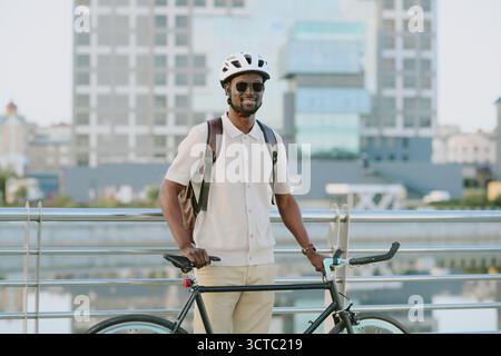 Giovane uomo nero adulto in piedi all'aperto tenendo in mano la bicicletta indossando il casco sorridente Foto Stock
