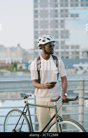 Giovane uomo nero adulto che indossa il casco che tiene lo smartphone accanto alla bicicletta Foto Stock