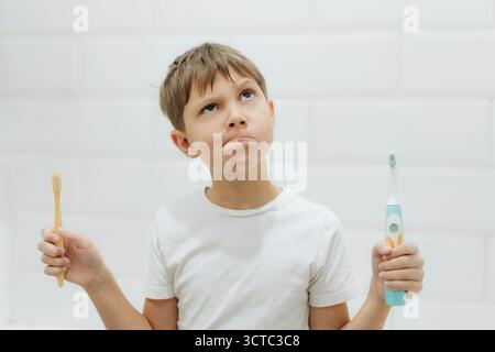 carino bambino di 8 anni che tiene in mano bambù e spazzolino elettrico per fare la scelta Foto Stock