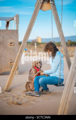 Donna seduta su un'altalena in un parco giochi sulla spiaggia di sabbia, tenuta per mano con il suo attento cane Weimaraner, simboleggiando il forte legame e l'amicizia Foto Stock
