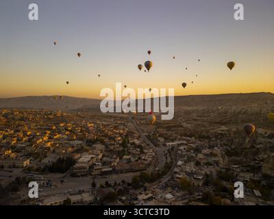 Veduta aerea di una moltitudine di mongolfiere che galleggiano serenamente sopra l'antico paesaggio, con la città di Goreme annidata sotto sotto la calda luce del sole, Gore Foto Stock
