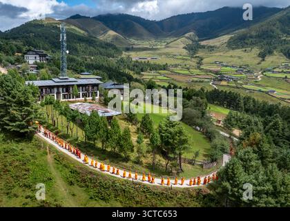 Vista aerea dei monaci in abiti di zafferano si snodano lungo un sentiero vicino a un edificio moderno sullo sfondo di montagne verdeggianti, Phobjikha, Wangdue Phodrang, B. Foto Stock
