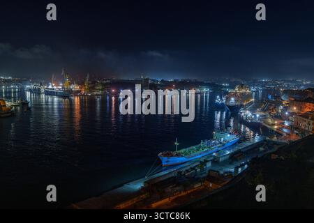 L'ampio panorama notturno mostra gru illuminate, banchine e navi ormeggiate che brillano attraverso il tranquillo porto. Foto Stock
