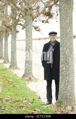 Uomo anziano, pensionato, con cappotto nero e cappellino si trova dietro un albero Foto Stock