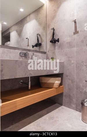 Moderno lavandino del bagno con lavabo in pietra naturale, ripiano in legno e specchio. Nelle vicinanze c'è un asciugacapelli che suggerisce servizi completi Foto Stock
