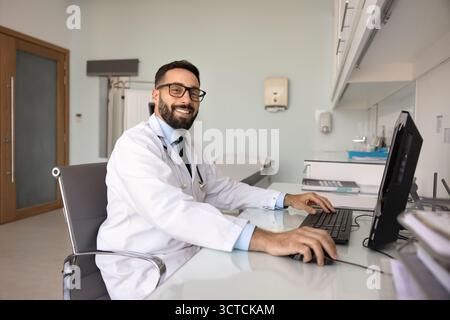 Ritratto di un medico ispanico sorridente seduto al suo tavolo sul posto di lavoro Foto Stock