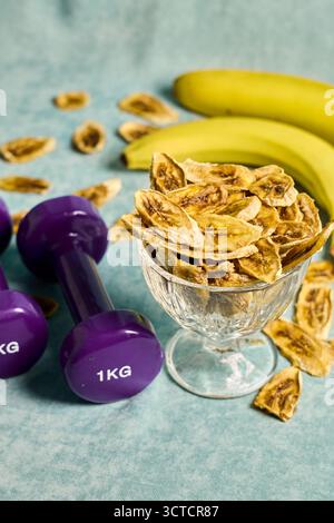 Concetto di stile di vita sano: Le patatine di banana in una ciotola di vetro con banane fresche e manubri viola su una superficie turchese testurizzata, che rappresenta il fitness Foto Stock