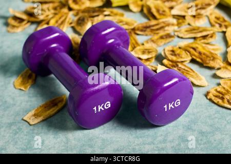 Immagine concettuale di scelte sane: Manubri viola e patatine di banana, che dimostrano l'equilibrio tra esercizio fisico e dieta per il benessere Foto Stock