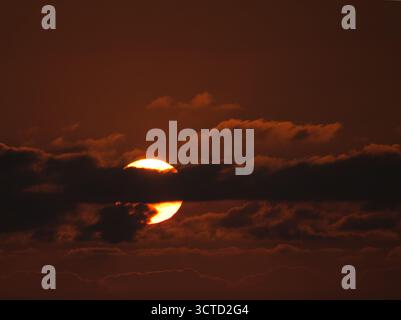 Burning Orange Sunset Over Ocean Horizon - intenso teleobiettivo primo piano del tramonto parzialmente nascosto da nuvole orizzontali scure che bagnano l'oceano Foto Stock