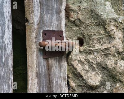 Serratura in metallo ossidato su legno e pietra antichi - primo piano di un bullone scorrevole pesantemente arrugginito contro tavole di legno intemperie e una struttura ruvida della parete in pietra. Foto Stock