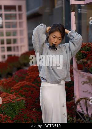 Bella giovane donna asiatica in piedi con un abito elegante, che usa una clip per capelli per legarsi i capelli. Scena urbana autunnale con vivace flusso di crisantemo rosso Foto Stock