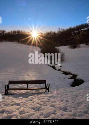 Sunburst Over Snowy Winter Landscape with Bench - panca solitaria in legno in un campo innevato incontaminato con bagliori di sole e effetto Starburst su un buio Foto Stock