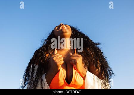 La giovane donna con lunghi capelli ricci chiude gli occhi e inclina la testa all'indietro, godendo il calore del sole sulla pelle Foto Stock