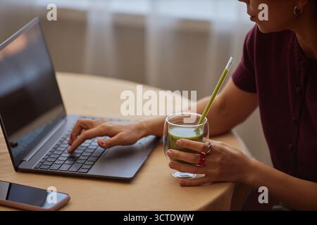 Giovane donna adulta che usa il laptop mentre beve il frullato verde Foto Stock