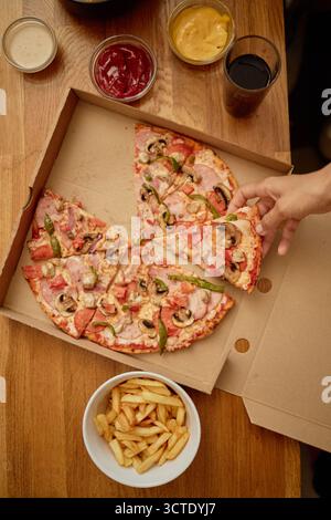 Giovane donna adulta che prende la fetta di pizza da Box con patatine fritte nelle vicinanze Foto Stock