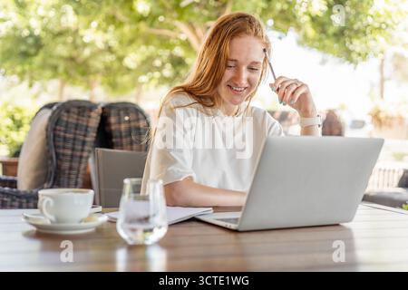 Giovane donna caucasica con lunghi capelli rossi sorride mentre lavora su un computer portatile in un bar all'aperto. Una tazza di caffè e un bicchiere d'acqua sono sul tavolo. Foto Stock