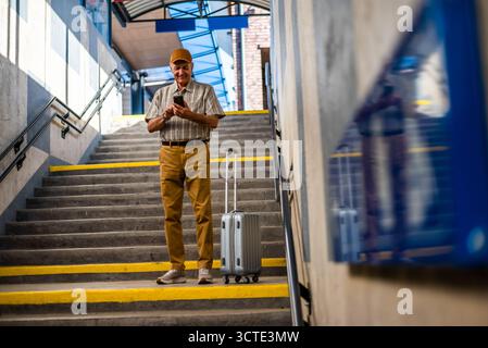 Un uomo anziano felice è in piedi alla stazione ferroviaria e invia messaggi sullo smartphone. Persone anziane in viaggio. Foto Stock
