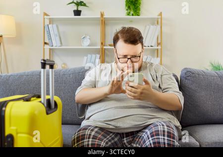 Uomo in sovrappeso che invia messaggi di testo sullo smartphone sul divano di casa, controlla la dieta e i piani di viaggio Foto Stock