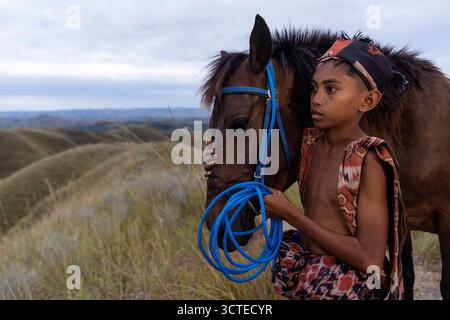 Ragazzo locale con abiti tradizionali e il suo cavallo su una collina erbosa nella parte orientale dell'isola di Sumba in Indonesia, ragazzo con tessuti tradizionali Foto Stock