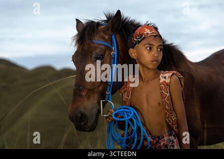 Ragazzo locale con abiti tradizionali e il suo cavallo su una collina erbosa nella parte orientale dell'isola di Sumba in Indonesia, ragazzo con tessuti tradizionali Foto Stock