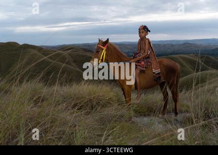 Ragazzo locale con abiti tradizionali e il suo cavallo su una collina erbosa nella parte orientale dell'isola di Sumba in Indonesia, ragazzo con tessuti tradizionali Foto Stock