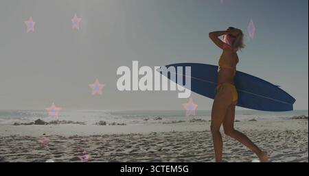 Surfista a piedi in bikini giallo che trasporta una tavola da surf blu sulla spiaggia con luce a stella e spazio per fotocopie Foto Stock