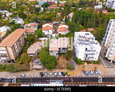 Rehovot, Israele - 11 settembre 2025: Veduta aerea di una vibrante scena urbana con case dal tetto in terracotta annidate tra vegetazione lussureggiante, contrastate da moderni grattacieli e auto parcheggiate ordinate. Foto Stock