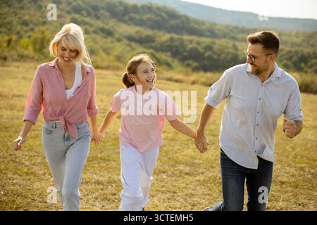 Una famiglia gioiosa, tra cui un padre, una madre e una figlia, cammina mano nella mano attraverso un prato soleggiato. Sorridono e si godono il pomeriggio Foto Stock