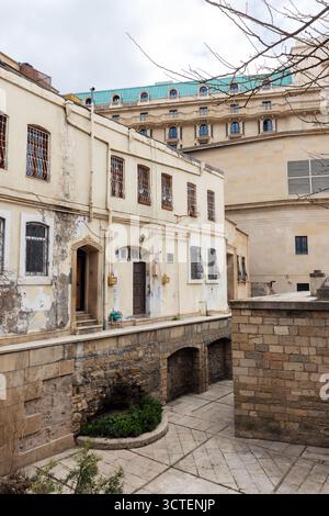 Un tranquillo cortile urbano incorniciato da edifici beige intemprati. Le finestre con barre di ferro e le porte ad arco si trovano accanto a un ponte di pietra. Baku, Azerbaigian. Vecchio Foto Stock