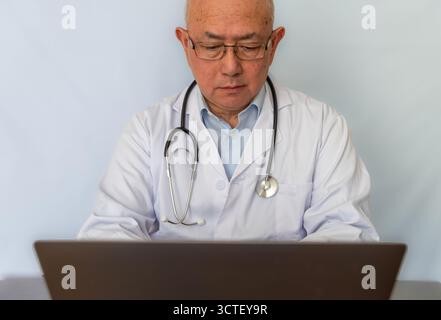 Un medico con un cappotto bianco con uno stetoscopio appeso al collo che lavora su un computer portatile su una scrivania. Foto Stock