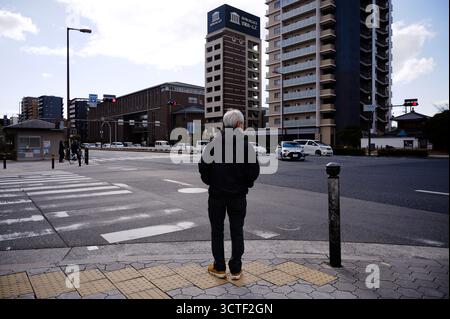 Un uomo anziano aspetta ad un incrocio su un tranquillo incrocio di Osaka, circondato da alti edifici residenziali Foto Stock
