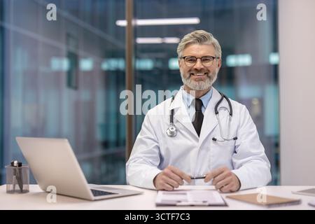 Medico maschile esperto in cappotto bianco con stetoscopio siede alla scrivania della clinica con computer portatile e documenti, sorridendo con sicurezza alla macchina fotografica trasmettendo cure mediche affidabili Foto Stock