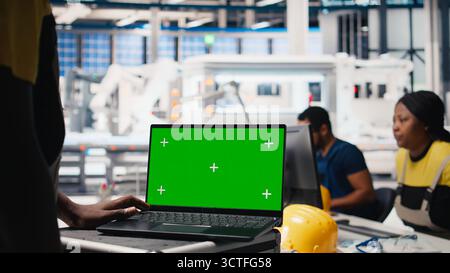 Le impostazioni del sistema di messa a punto di un esperto di impianti fotovoltaici su un PC con schermo verde. Il responsabile lavora su un laptop presso l'impianto di produzione di pannelli solari per la configurazione delle linee di assemblaggio. Telecamera B.. Foto Stock