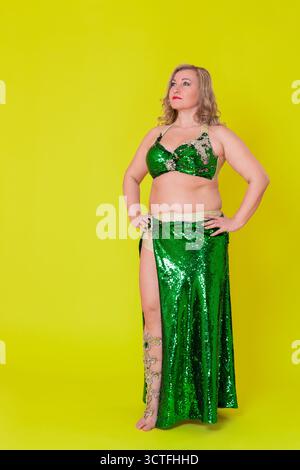 Modella caucasica vestita con abito da ballo - top con paillettes verde e gonna lunga con spacco a piedi nudi su tutta la lunghezza contro ba gialla Foto Stock