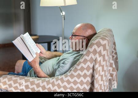 Un uomo anziano gode di un momento tranquillo leggendo un libro seduto su una sedia confortevole. La luce soffusa emessa da una lampada vicina aggiunge calore all'atmosfera rilassante. Foto Stock