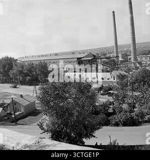Un paesaggio industriale a Sloviansk, RSS Ucraina, durante gli anni '1970 Questa fotografia d'archivio mostra una grande fabbrica o un impianto di lavorazione, probabilmente un complesso di elevatore di grano, che domina il distretto. Due massicce aree affumicate si innalzano in alto sopra l'edificio principale, simboleggiando il potere industriale della regione del Donbas. In primo piano, le strade residenziali con auto come la classica Moskvitch-412 coesistono con la zona industriale. Questa immagine cattura la tipica pianificazione urbana dell'era sovietica, dove la vita industriale e residenziale era strettamente intrecciata Foto Stock