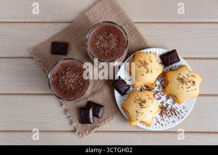 Deliziosi biscotti fatti in casa serviti con budino al cioccolato in tazze di vetro, ricoperti di cioccolato fondente grattugiato. Accogliente composizione di dessert su un tavolo di legno Foto Stock